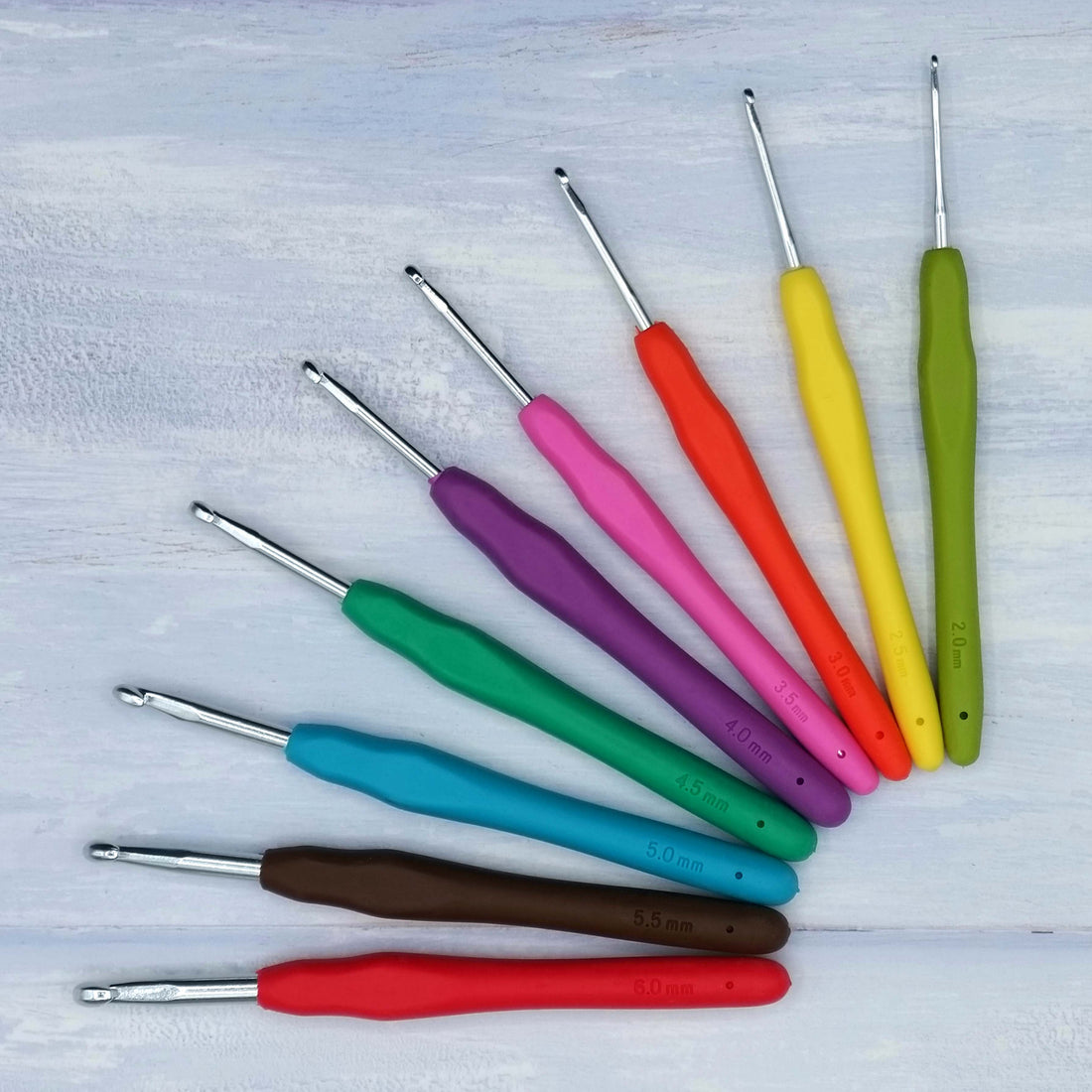 A fan of colourful crochet hooks with soft grip handles in various sizes, laid out on a light wooden background